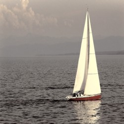 Sailboat with red hull glides across calm waters on a sunny serene day.