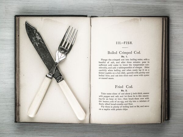 Old cookery book open with an old fork spoon and table knife by Assaf Frank