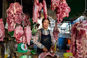 Woman butcher at market stall with hanging raw meat.