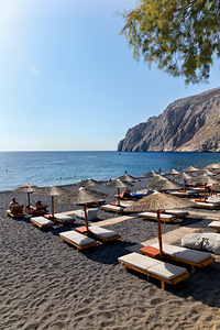Sunny black sand beach with umbrellas sunbathers and a cliff.
