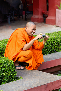 Smiling monk in orange robes takes photo with digital camera.