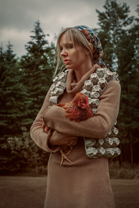 Young blonde woman with a scarf on her head wearing a knitted dress and a down vest made of egg cartons holding a brown hen in her arms near a coniferous forest