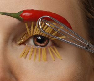 Close-up of a womans brown eye with a whisk as a mascara applicator and spaghetti pastas as eyelashes her eyebrow made of a red hot pepper
