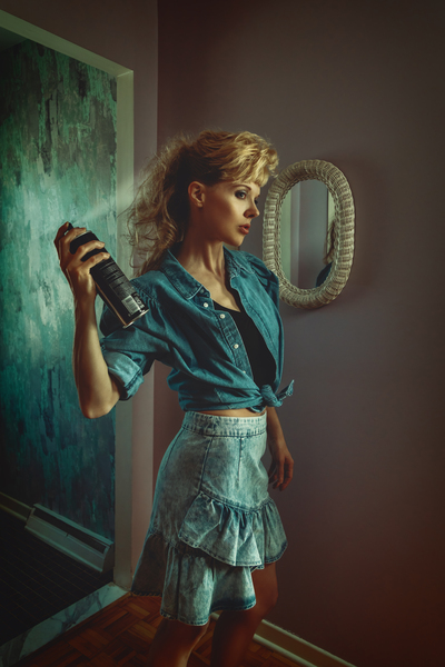 vintage lifestyle portrait of a young blonde woman in a late 80s early 90s interior decor wearing a denim outfit and styling her teased hair and bangs with hairspray by Artmood Visualz