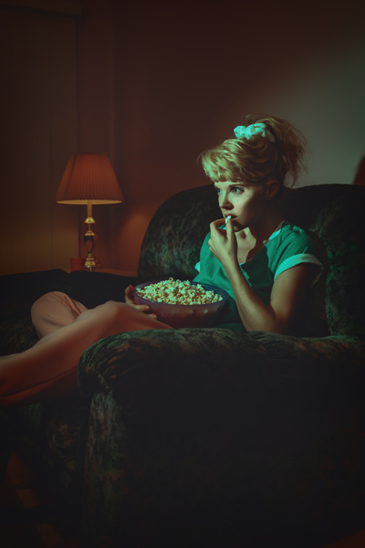 vintage lifestyle portrait of a young blonde woman in a late 80s early 90s interior decor eating popcorn andrelaxing on a sofa while watching television in the dark by Artmood Visualz