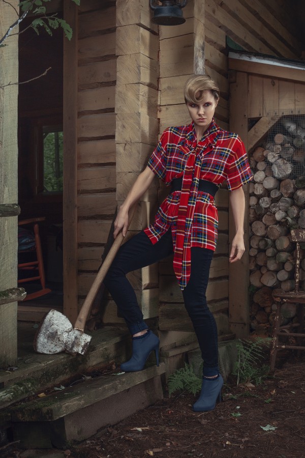 Beautiful young blonde woman with a curl in her hair wearing a long plaid shirt standing on the stairs of a rustic wooden shack an antique axe leaning against her leg by Artmood Visualz