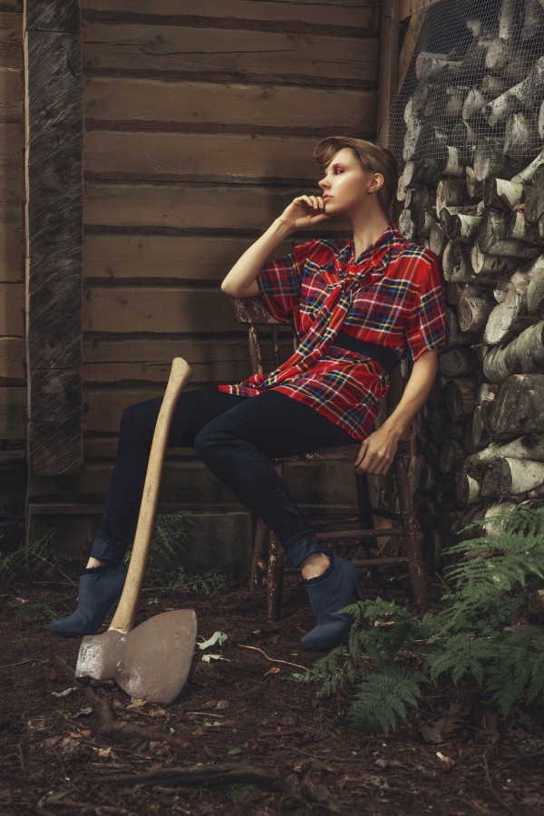 Young beautiful woman with short hair wearing a long plaid shirt sitting on a chair outside of a shack with an antique axe leaning against her leg by Artmood Visualz