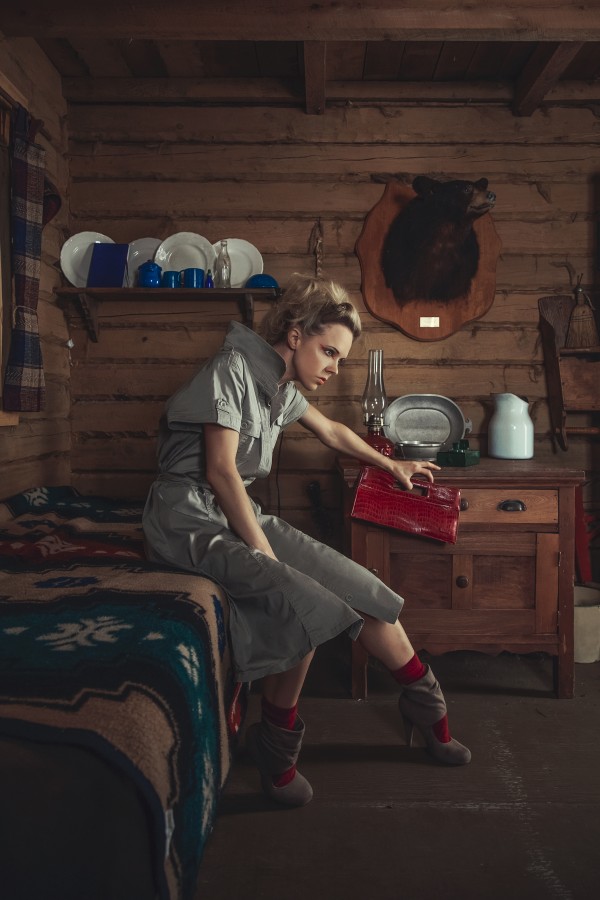Side view of a beautiful young blonde woman in a beige dress holding a red handbag sitting on the bed of a rustic wooden cabin by Artmood Visualz