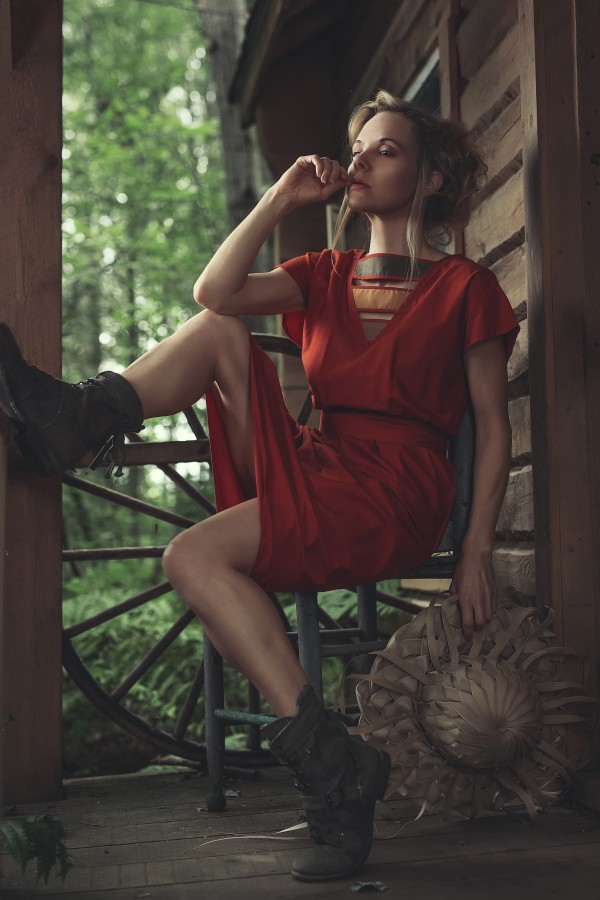 Young beautiful woman in a red casual dress relaxing while sitting on the balcony of an old shack holding a braided palm sun hat by Artmood Visualz