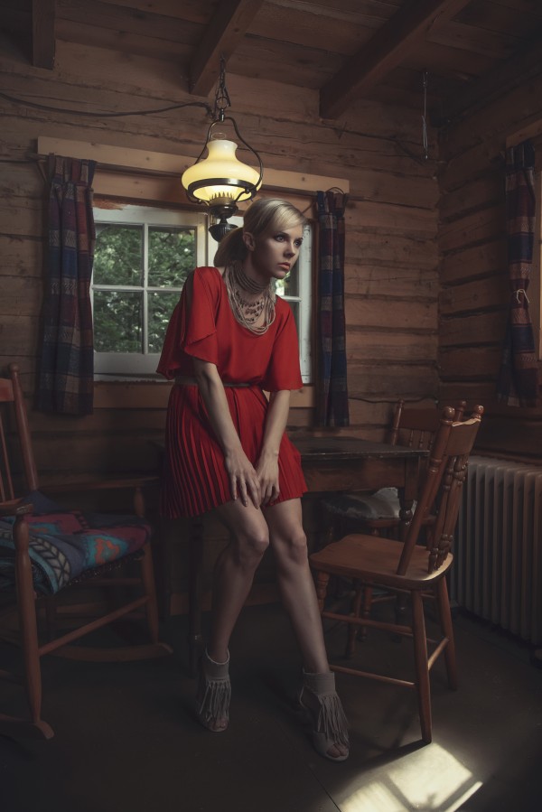 Beautiful young blonde woman in a dark orange dress sitting on the table of a rustic wooden shack by Artmood Visualz