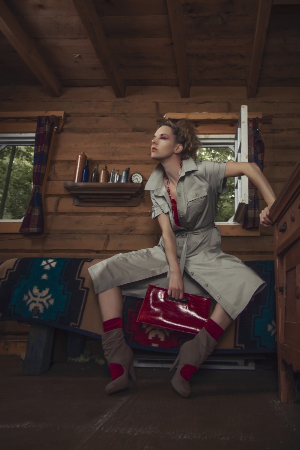Young blonde woman in a beige dress holding a red handbag sitting on the bed of a rustic wooden cabin by Artmood Visualz