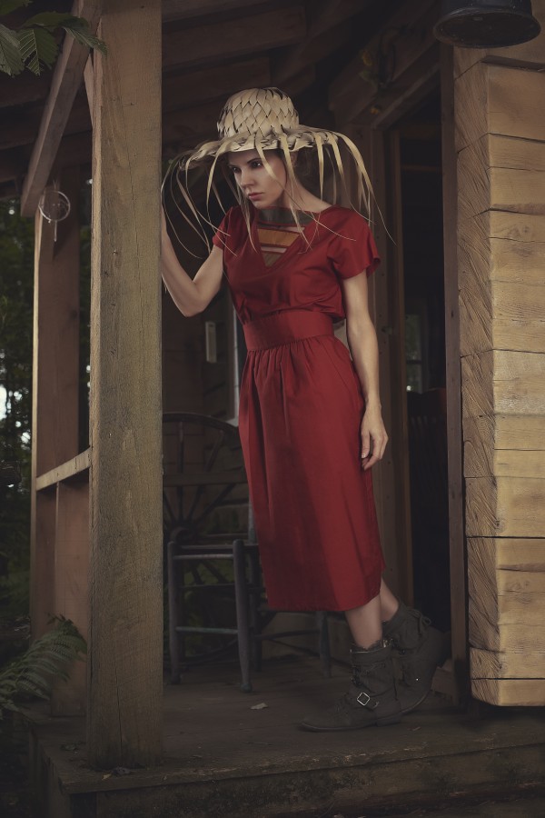 Young beautiful woman in a red casual dress wearing a braided palm sun hat standing on the balcony of an old shack by Artmood Visualz