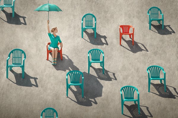 Young blonde woman holding up a turquoise umbrella on a beach sitting through many red and teal chairs in the sand on a sunny day Print