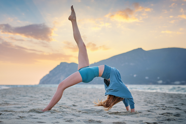 Woman practicing Eka Pada Urdhva Dhanurasana on sandy beach at sunset by Dmitry Rukhlenko