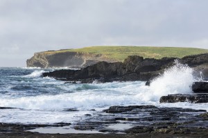 Waves crashing into rocky coast with large grassy hill and cliffs in background; Kilkee, County Clare, Ireland