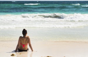 Young woman sitting on sandy beach in water looking out at waves coming in and blue sky; Akumal, Quintana Roo, Mexico