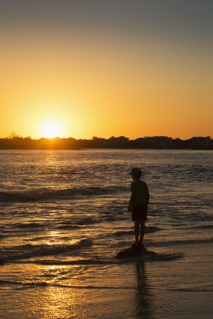 Silhouette of a young boy standing at the water's edge at sunset; Caloundra, Queensland, Australia