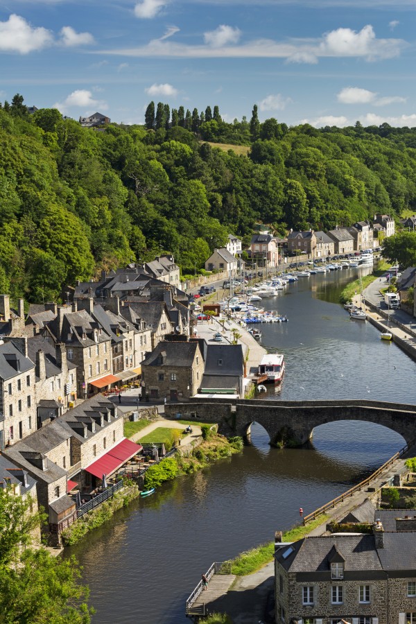 Treed valley riverside town, stone bridge and boats in the harbour with blue sky and clouds; Dinan, Brittany, France by PacificStock