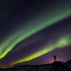 Colourful Aurora borealis over a man with arms outstretched silhouetted against light pollution from nearby Fort Greely; Alaska, United States of America