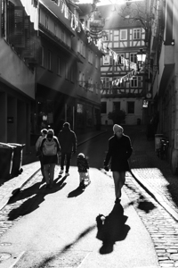 Black and white street photo of an older man walking through a sunlit alley 