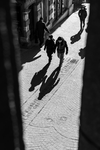 Black and white street photo of people walking down a road in the city seen through a fence
