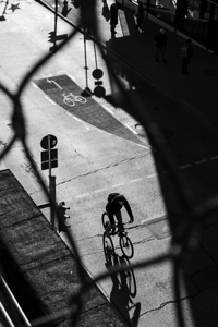Black and white street photo of a bicyclist on a road in the city seen through a fence