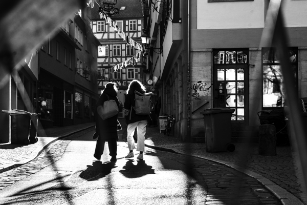 Black and white street photo of people walking down the road seen through the spokes of a bicycle Print