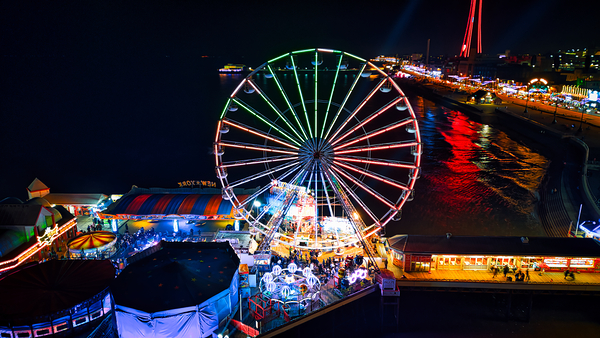 Colorful night view of a vibrant amusement park with a Ferris wheel and bright lights reflecting on water in Backpool England. by Vasile Jechiu