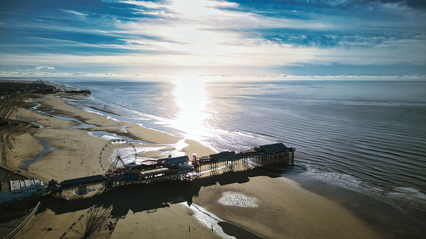 Aerial view of a sunlit beach with a pier extending into calm waters casting shadows on the sand in Backpool England. by Vasile Jechiu