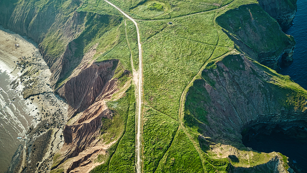 Aerial view of a winding road cutting through lush green hills with coastal cliffs and ocean in the background in Filey England. by Vasile Jechiu