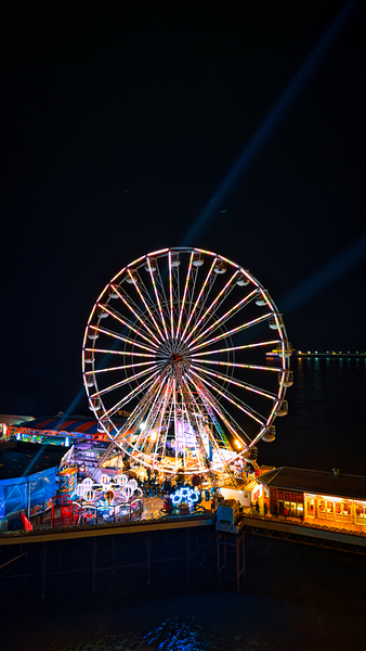 Illuminated Ferris wheel at night in an amusement park with vibrant colors and festive lights in Backpool England. by Vasile Jechiu