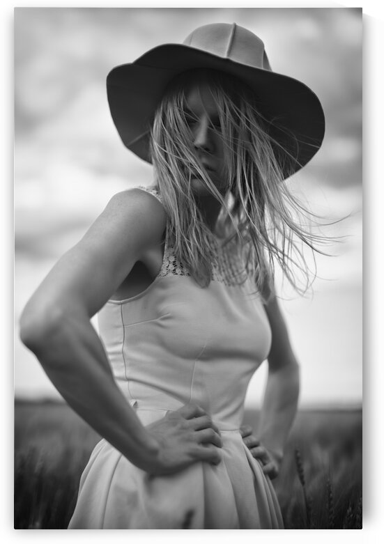 Black and white portrait of a beautiful young woman wearing a summer dress and a sun hat standing outdoors in a wheat field on a cloudy day by Artmood Visualz