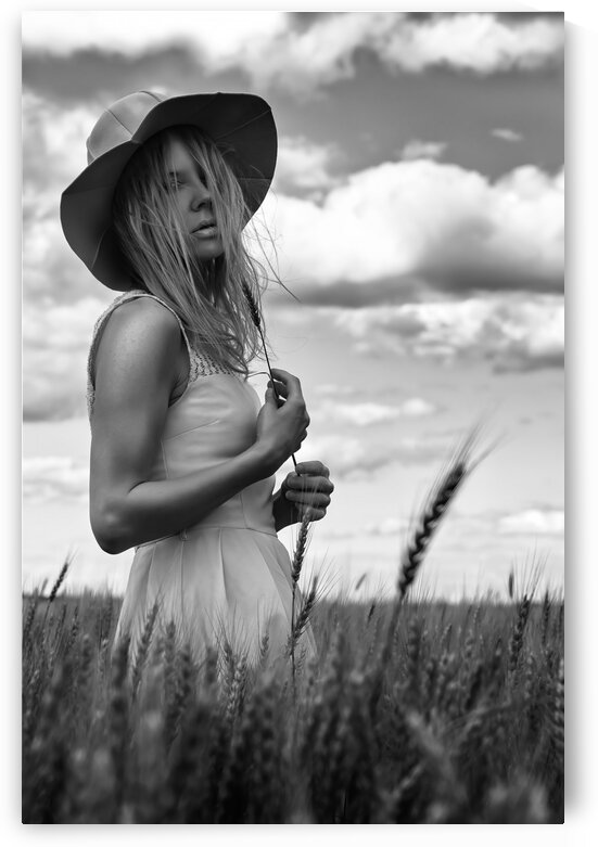 Black and white portrait of a beautiful young woman wearing a summer dress and a sun hat standing outdoors in a wheat field on a cloudy day by Artmood Visualz