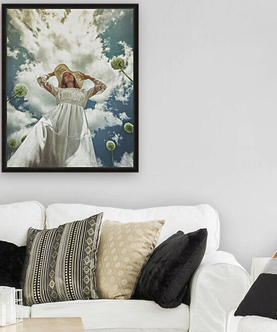 Ground angle view of a young blonde woman in a white dress with a white hat surrounded by dandelions under a blue sky with white clouds by Artmood Visualz