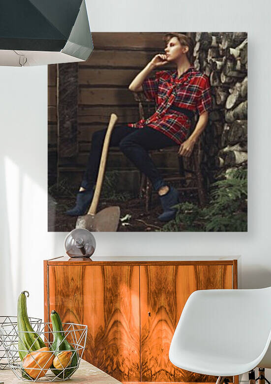Young beautiful woman with short hair wearing a long plaid shirt sitting on a chair outside of a shack with an antique axe leaning against her leg by Artmood Visualz