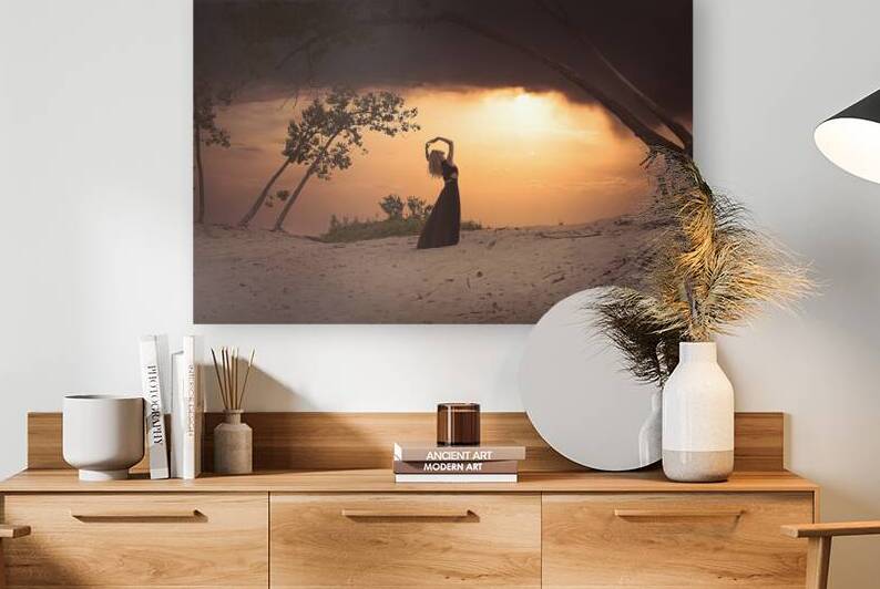 Blonde woman standing on a beach facing the setting sun with her arms raised and wearing a long black dress by Artmood Visualz